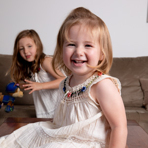 Young girl indoors smiling, her sister playing in background.