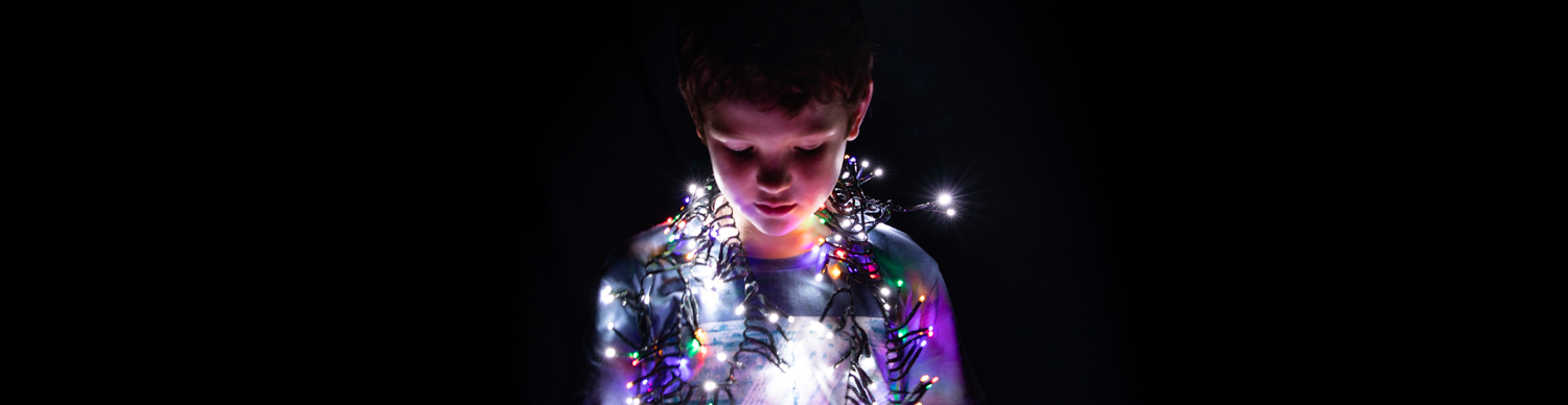 a young boy in the dark with fairy lights draped around his shoulders casting their glow on his face
